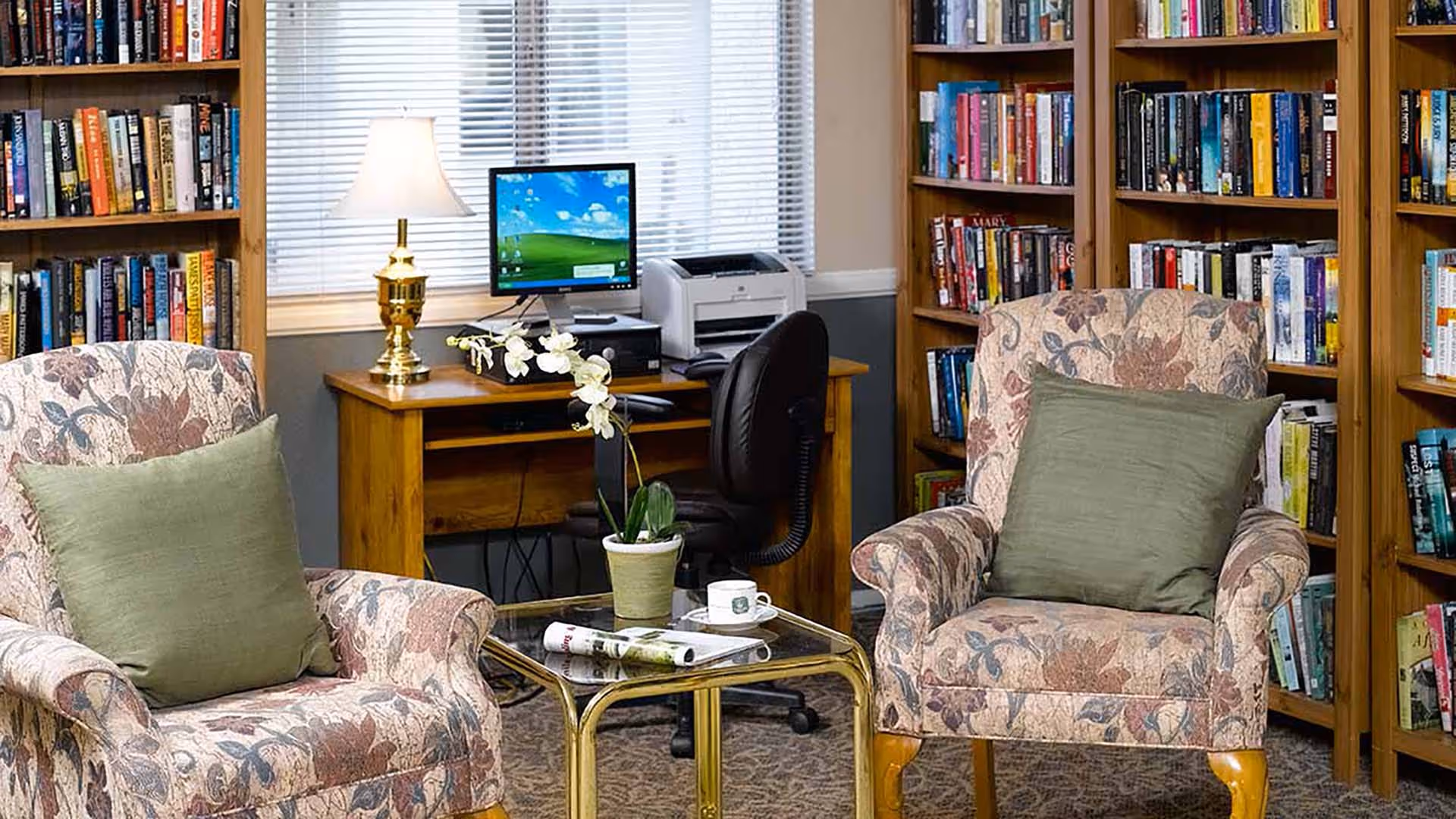 A cozy library or reading room with two floral upholstered armchairs each with a green cushion, a glass-top coffee table with a potted orchid, a newspaper, and a cup and saucer. Behind the chairs are wooden bookshelves filled with books, and a wooden desk with a computer, printer, and a brass table lamp in front of a window with blinds.