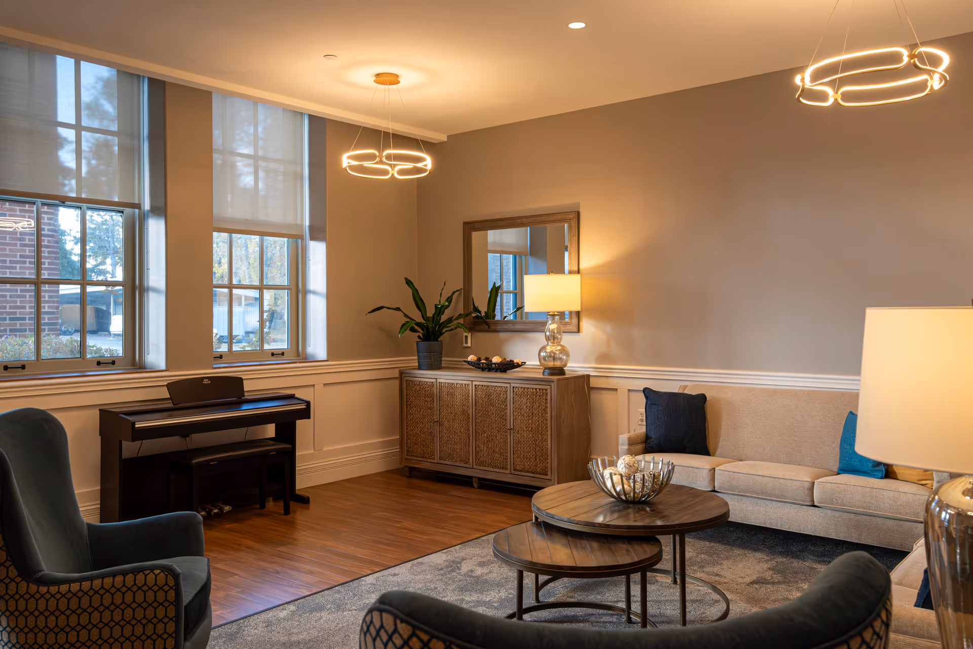 A warmly lit living room with a sofa, armchairs, nested coffee tables, a piano, sideboard with a lamp and large windows.