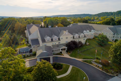 Aerial view of Simpson Meadows Retirement Community, showing a large multi-wing building surrounded by green lawns, trees, and a curved driveway leading to the entrance. The landscape includes rolling hills and a partly cloudy sky in the background.
