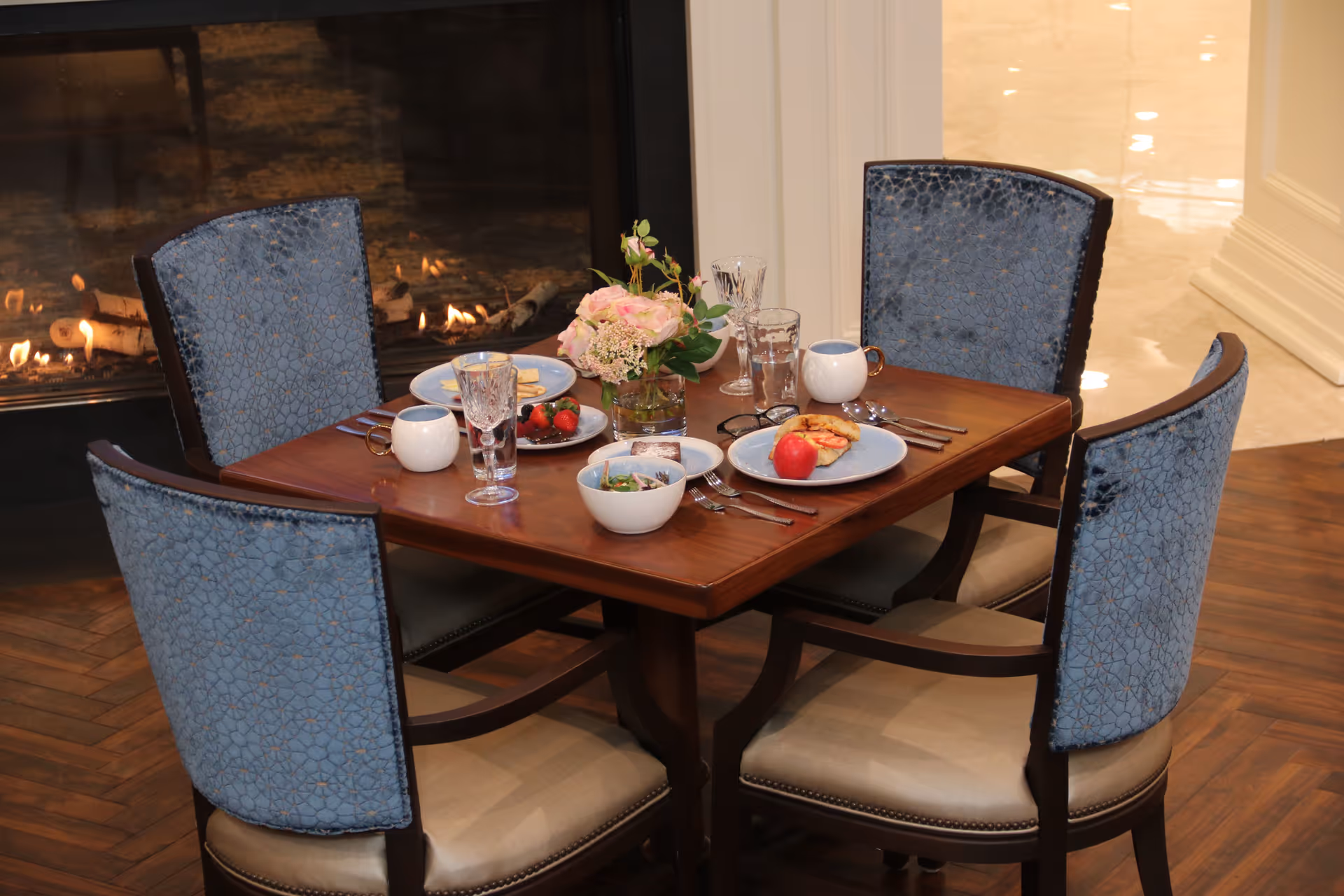 Small wooden dining table set for four with blue-upholstered chairs, place settings, a vase of flowers, and a lit fireplace in the background.