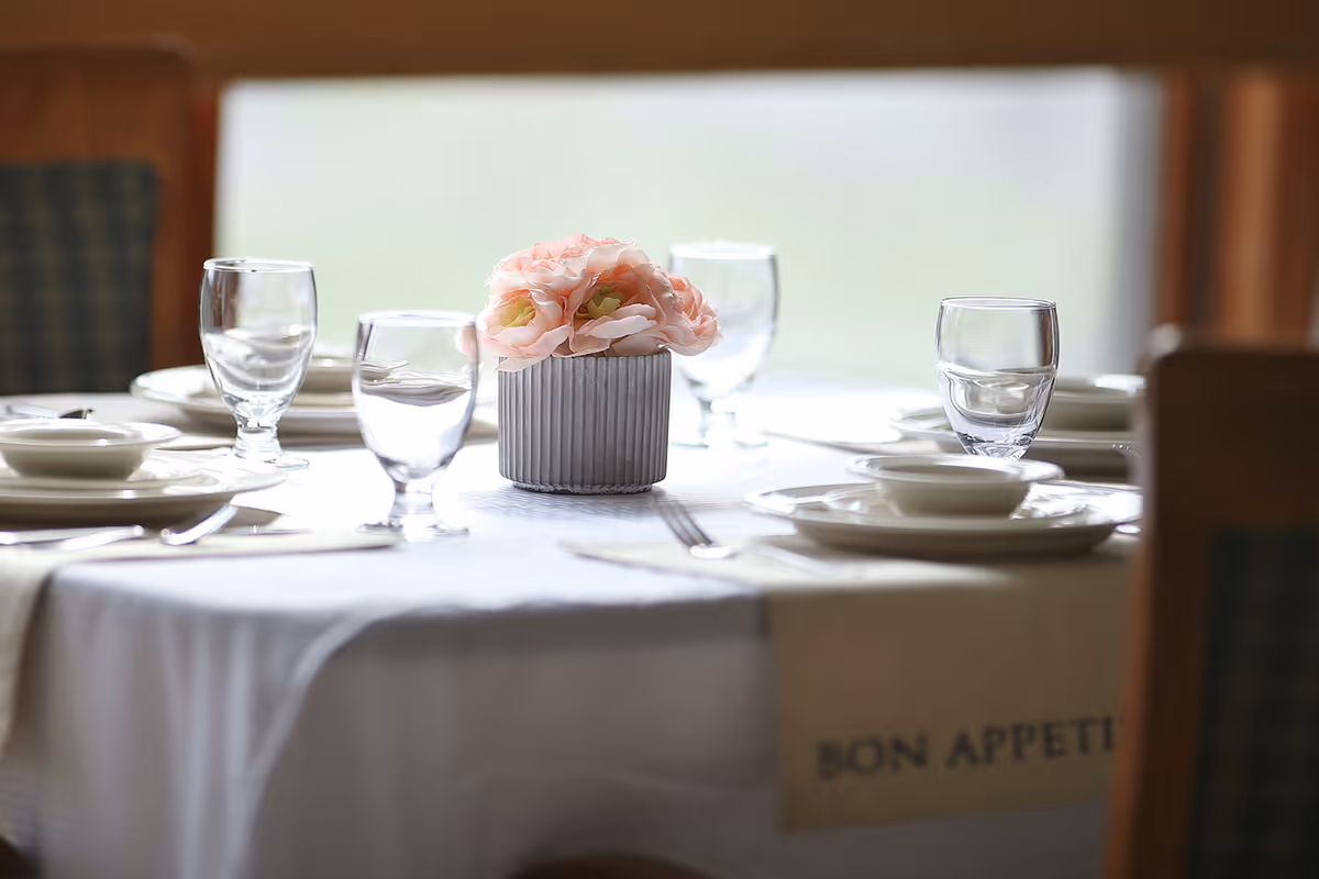 A round dining table set with clear water glasses, white plates, bowls, and silverware. A small vase with light pink flowers is placed in the center of the table. The table is covered with a white tablecloth and a beige cloth with the words 'BON APPETIT' partially visible. Chairs with patterned upholstery surround the table.