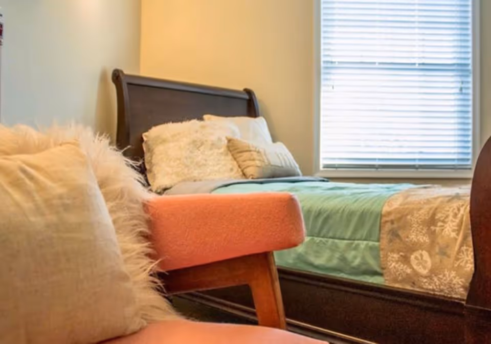 A cozy bedroom featuring a dark wooden bed with light-colored pillows and a teal blanket, next to a window with white blinds. In the foreground, there is a coral upholstered chair with a beige fluffy pillow.