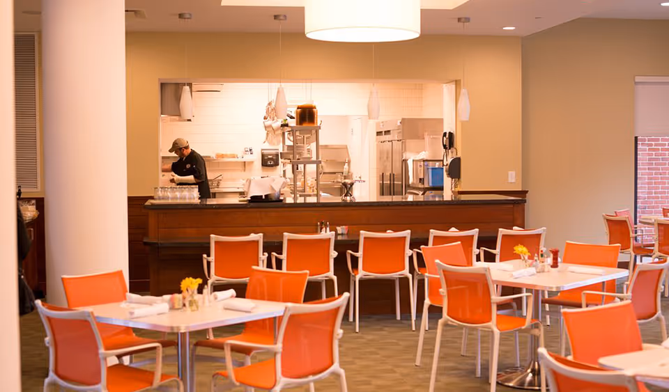 Dining room with orange chairs and white tables facing an open serving counter where a staff member works in the kitchen.