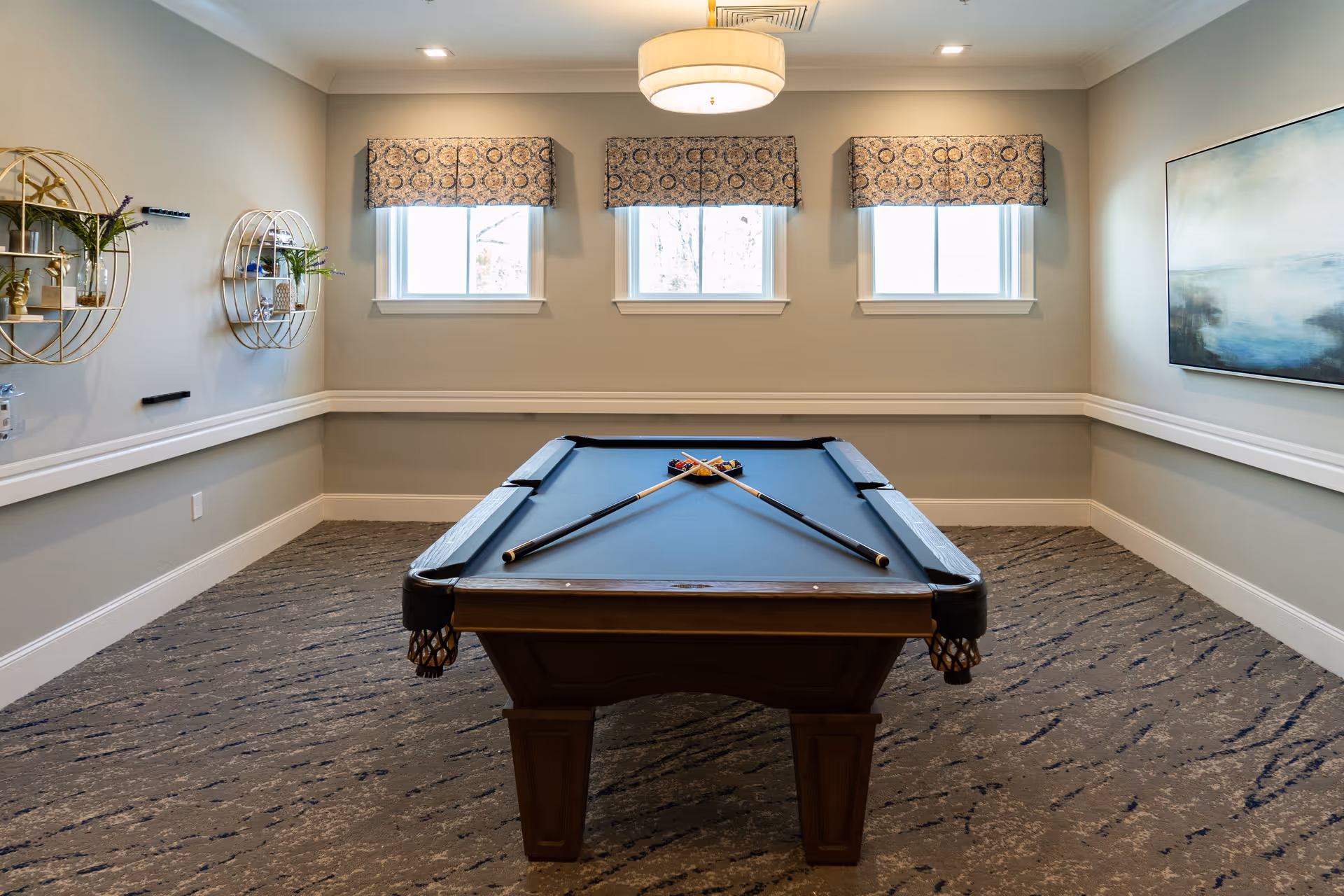 A well-lit game room with a pool table centered in the room. The pool table has two crossed cues and a rack of billiard balls. The room features three windows with patterned valances, two circular wall shelves with decorative items and plants on the left wall, and a large abstract painting on the right wall. The carpet has a textured pattern, and the walls are painted a light neutral color.