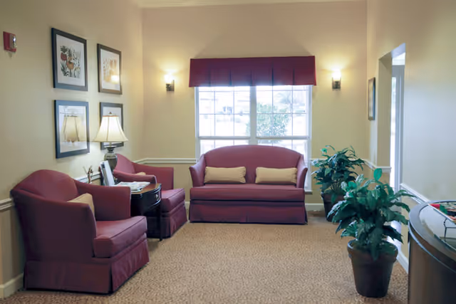 A cozy sitting area with two maroon armchairs and a matching loveseat with beige cushions. There is a side table with a lamp and framed pictures on the wall. Two potted plants are placed on the right side near a window with a maroon valance, and soft wall sconces provide lighting.