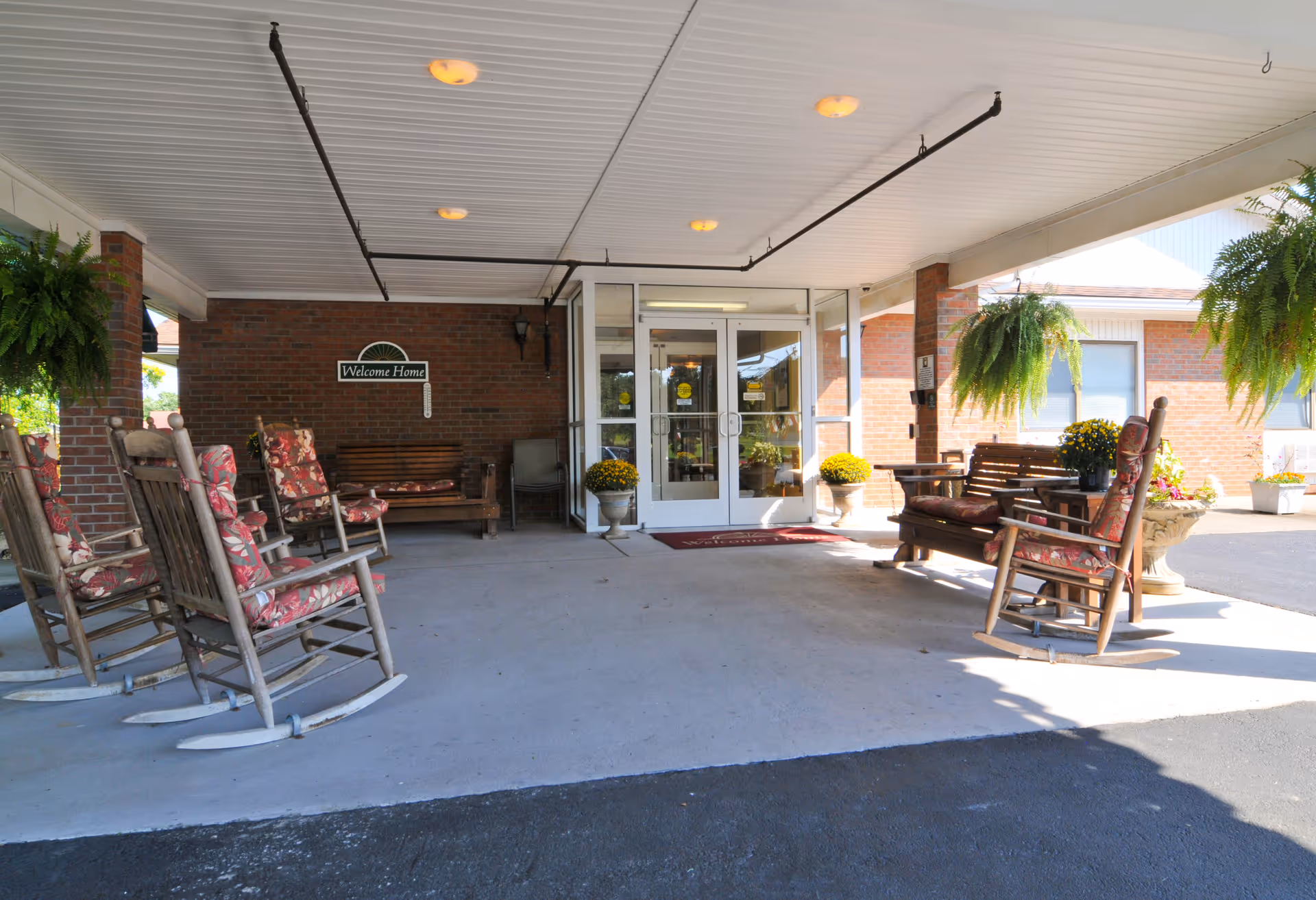 Covered entrance area of a senior living facility with several wooden rocking chairs and benches with floral cushions arranged on either side. Hanging green plants and potted yellow flowers decorate the space. A brick wall with a 'Welcome Home' sign is visible near the glass double doors leading inside.