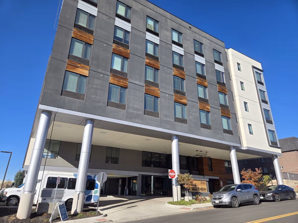 The Belleview Senior Living multi-story building front with a covered entrance, pillars and parked cars under a clear blue sky.
