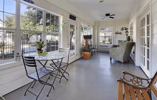 Bright enclosed sunroom with a bistro table and chairs, an armchair, and large windows overlooking a fenced yard.