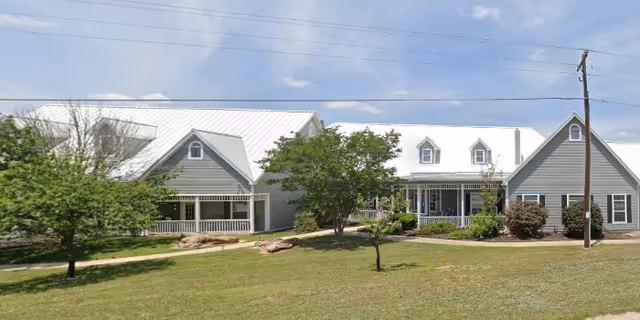 Exterior view of a senior living facility named Arbor House Fredericksburg featuring two connected gray buildings with white roofs, surrounded by green grass, trees, and shrubs under a partly cloudy sky.