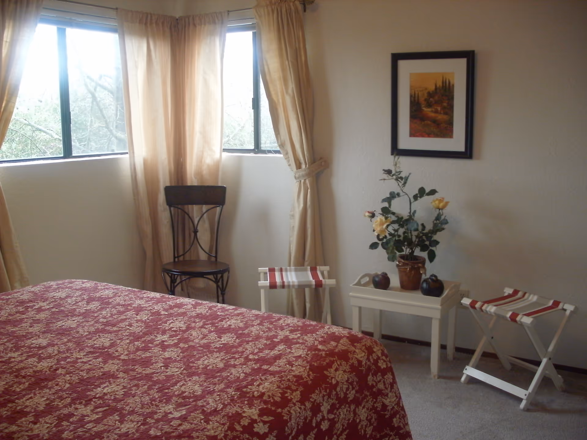 Bedroom with a red floral bedspread, corner windows with curtains, a chair, a small table holding a potted plant and decorative items, and two folding stools.