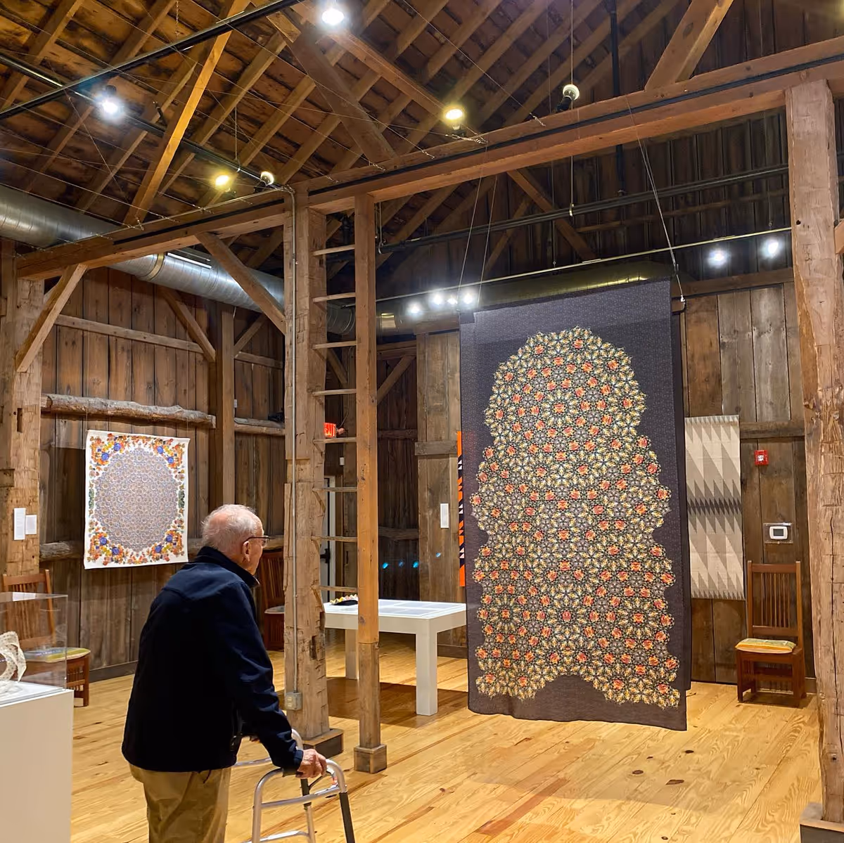 An elderly man using a walker is viewing intricate textile art pieces displayed in a rustic wooden gallery with exposed beams and wooden floors.
