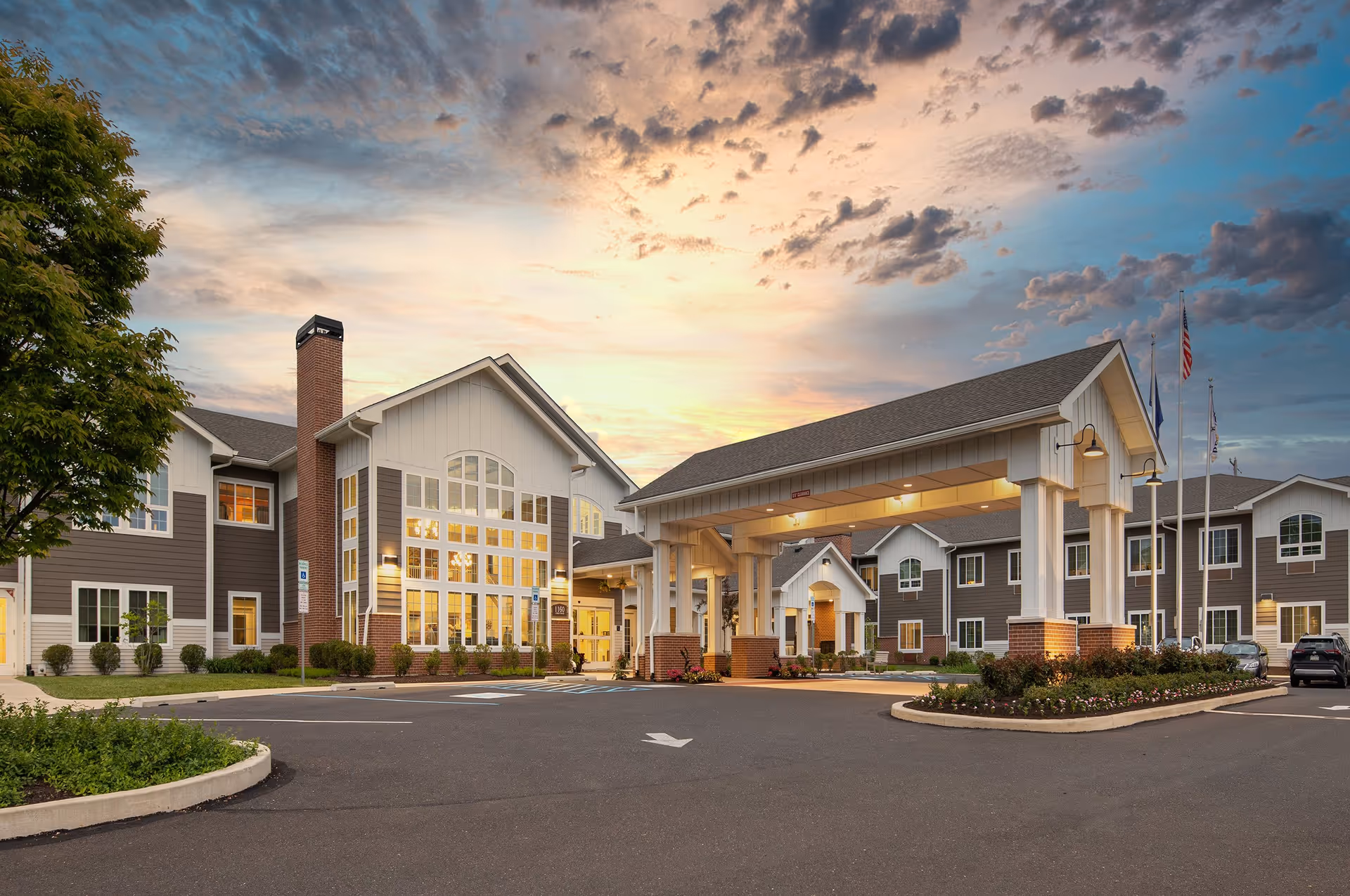 Exterior view of The Province of Southampton senior living facility at sunset, showing a large building with multiple windows, a covered entrance supported by white columns with brick bases, landscaped greenery, and a parking area with a few cars.