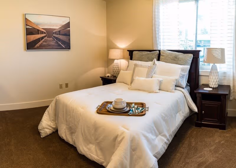 A cozy bedroom with a neatly made bed featuring white bedding and multiple pillows. There is a wooden tray with a cup and saucer on the bed. Two dark wooden nightstands flank the bed, each with a white patterned lamp. A large window with white blinds allows natural light into the room. A framed picture of a wooden boardwalk leading to the beach hangs on the wall.
