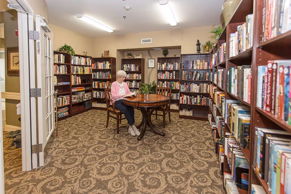 A cozy library room with carpeted floor and several wooden bookshelves filled with books. An elderly woman wearing a pink cardigan and white shoes is sitting at a round wooden table in the center, reading a book. The room is well-lit with fluorescent ceiling lights and decorated with small plants on the shelves and table.