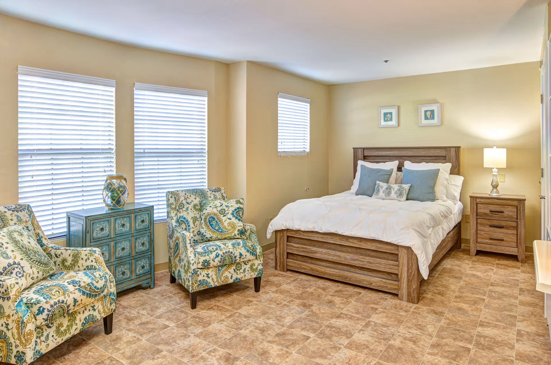 A bright bedroom with a wooden bed frame, white bedding, and multiple pillows. Two patterned armchairs with blue and green designs are placed near three windows with white blinds. A small blue chest of drawers sits between the chairs, and a wooden nightstand with a lamp is beside the bed. The floor has a light brown tile pattern, and two framed pictures hang above the bed.