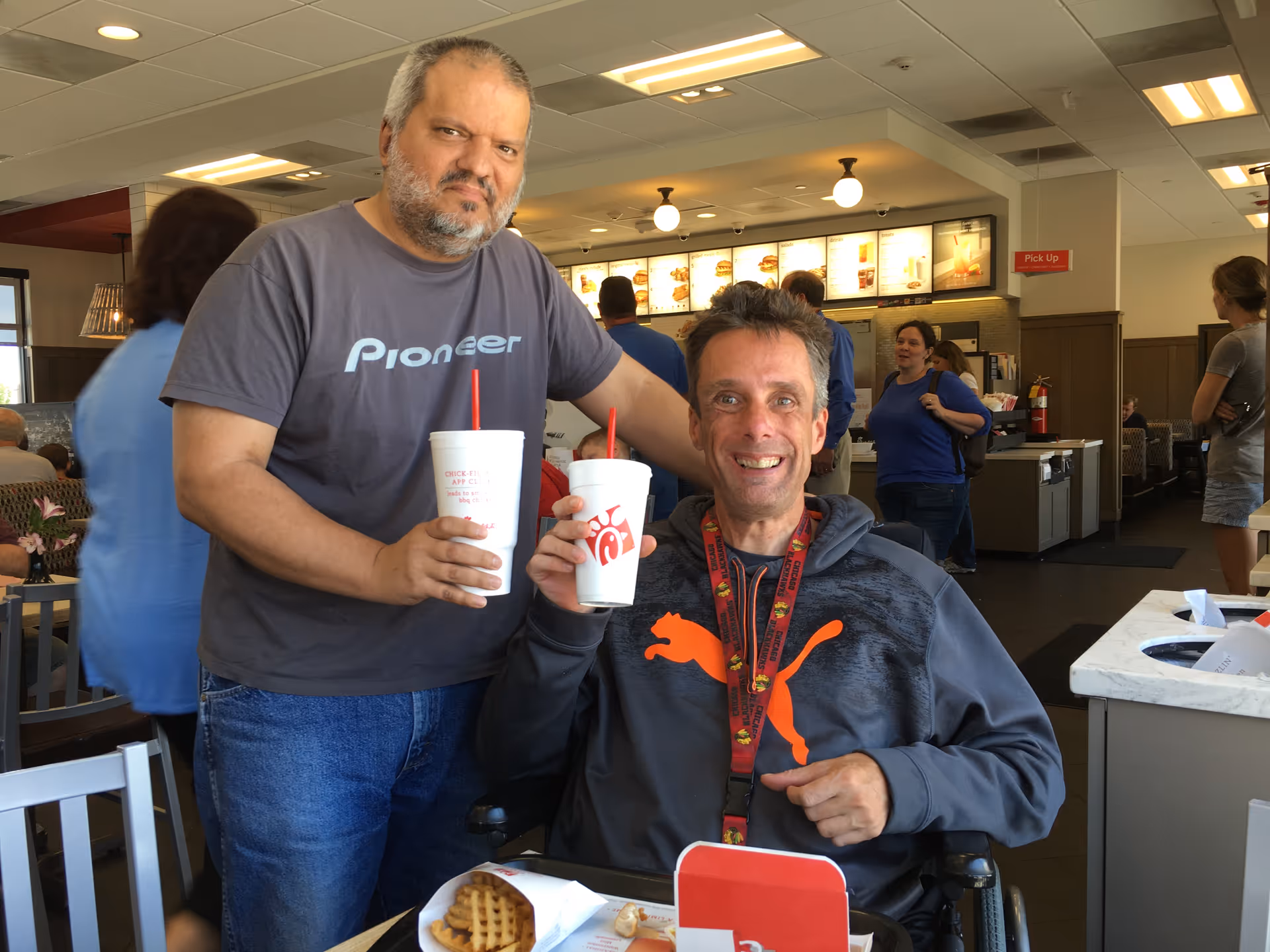 Two men inside a fast food restaurant, one standing and holding a drink cup, the other sitting in a wheelchair also holding a drink cup. The man sitting is smiling and wearing a gray Puma hoodie. The background shows other customers and a menu board with food items.