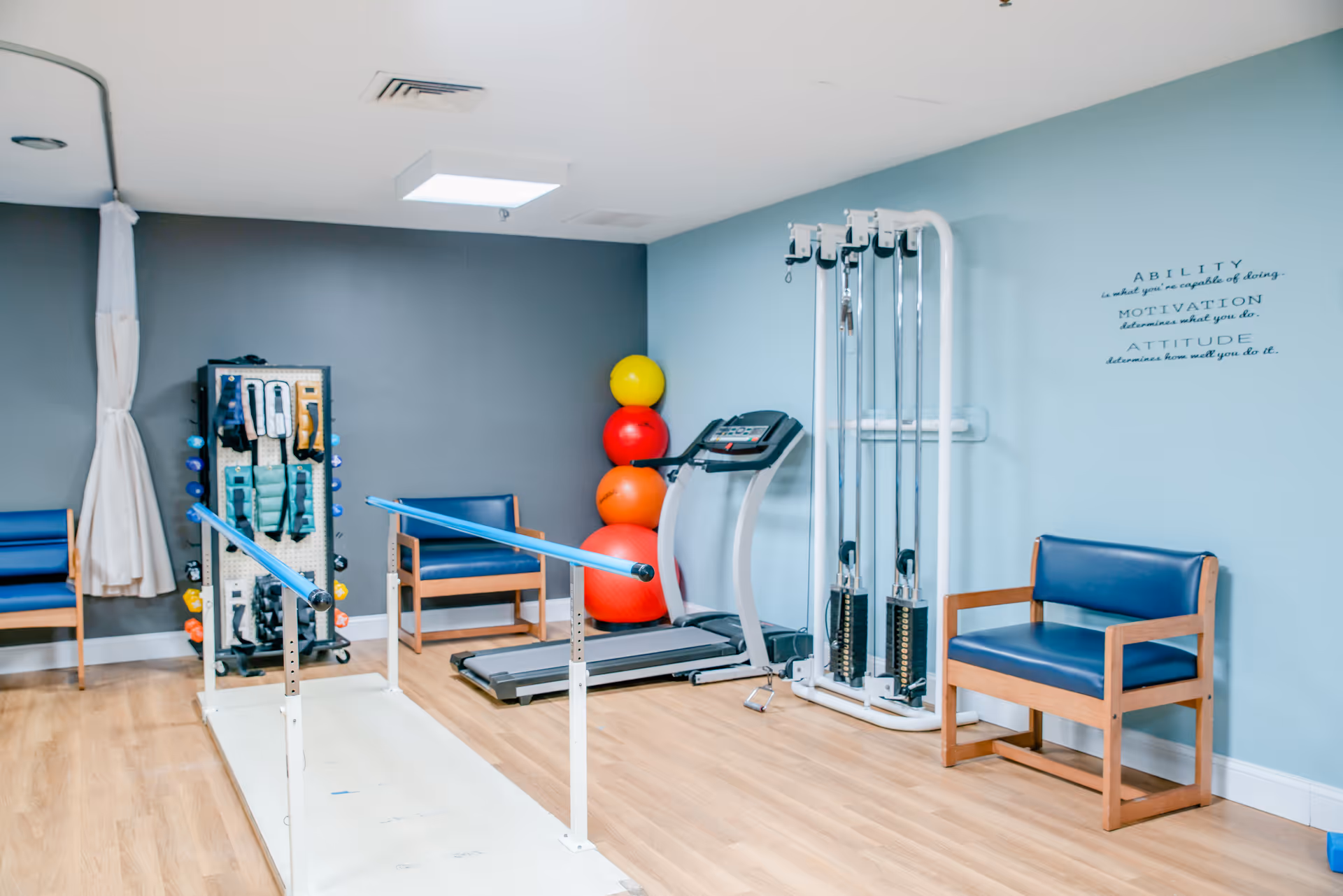 A physical therapy or exercise room with parallel bars for walking support, a treadmill, exercise balls stacked in the corner, weight machines, and two blue cushioned wooden chairs against the walls. The walls are painted light blue and gray, and there is a motivational quote on the wall.