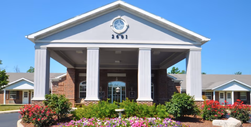 Front exterior view of a brick building with a white portico supported by four large columns, surrounded by landscaped flower beds with colorful flowers and green shrubs under a clear blue sky.