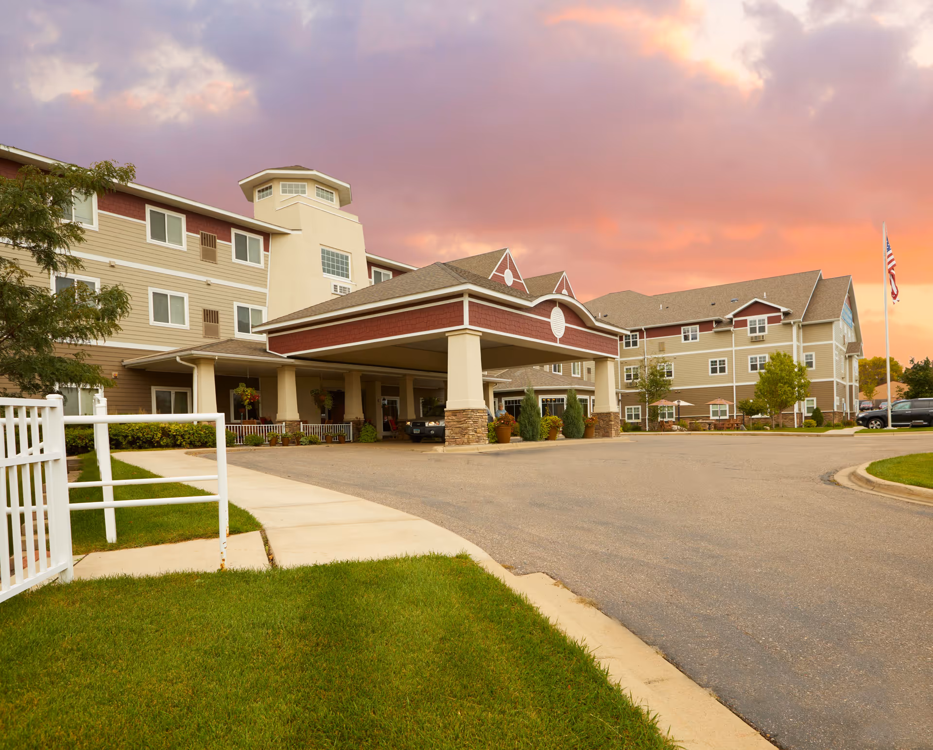 Front entrance of a multi-story senior living building with a covered porte-cochère, landscaped lawn, and a colorful sunset sky.