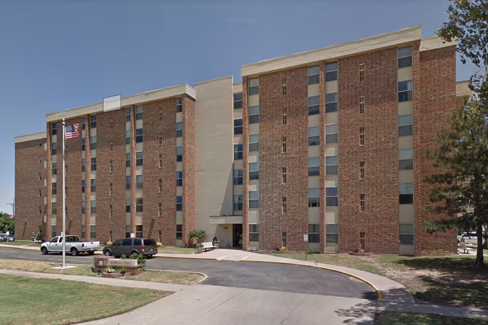 Exterior view of a multi-story brick building with several windows, an entrance with a small awning, a flagpole with an American flag, and a few parked vehicles in front. The sky is clear and blue.