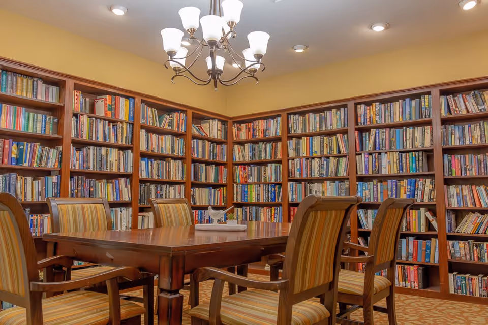 A cozy library room with wooden bookshelves filled with books lining the walls. In the center, there is a wooden table surrounded by six striped upholstered chairs. A chandelier with multiple lights hangs from the ceiling, illuminating the room with warm lighting.