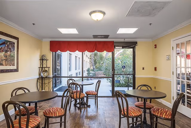 Interior view of a small dining area with three round tables and several chairs with striped cushions. The room has yellow walls with white trim, a framed painting on the left wall, and a glass door leading to an outdoor patio with greenery and a bench. A red valance is above the glass door.