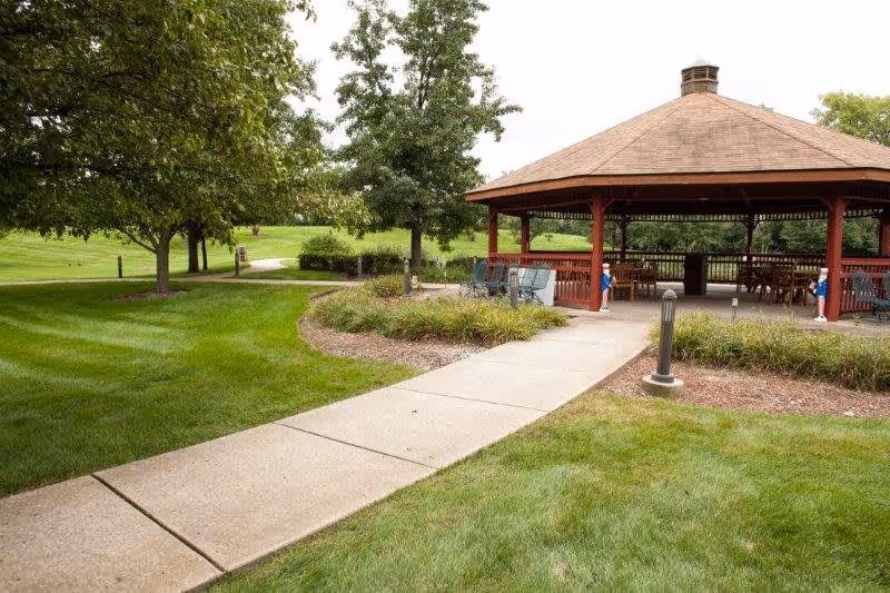 A paved walkway leads to a wooden gazebo with a brown roof in a green outdoor area with trees and well-maintained grass.