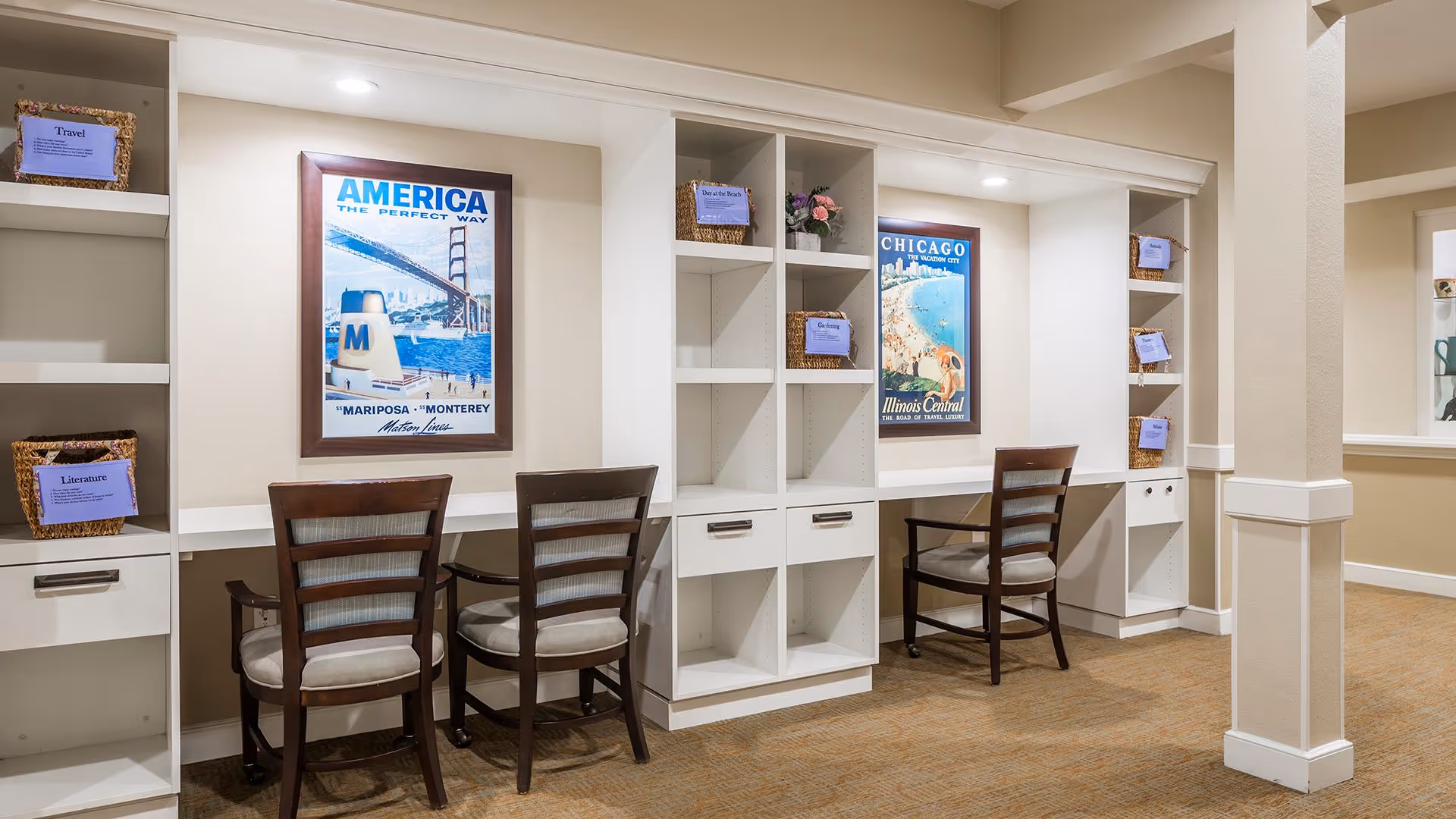 A well-lit interior space with built-in white shelving units and desks. There are three wooden chairs with cushioned seats placed in front of the desks. The shelves contain wicker baskets labeled with categories such as Travel, Literature, Day at the Beach, and Gardening. Two framed vintage travel posters are mounted on the wall above the desks, one featuring America and the other Chicago. The room has beige walls and carpeted flooring.