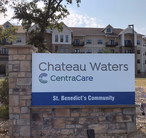 A stone-mounted entrance sign reading 'Chateau Waters CentraCare St. Benedict's Community' with a multi-story residential building in the background.