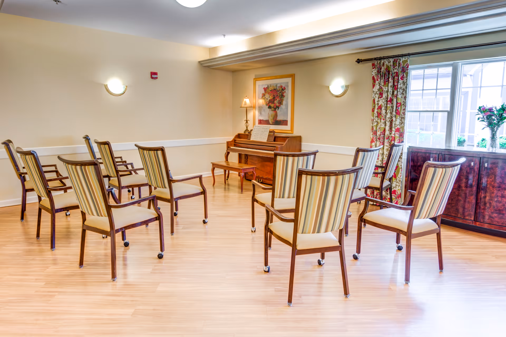 A bright room with wooden flooring featuring a small upright piano against the wall with a bench in front. Surrounding the piano are several wooden chairs with striped upholstery arranged in a semi-circle. There is a framed floral painting above the piano, a small table lamp, and a large window with floral curtains letting in natural light. A dark wooden cabinet with a vase of flowers is positioned near the window.