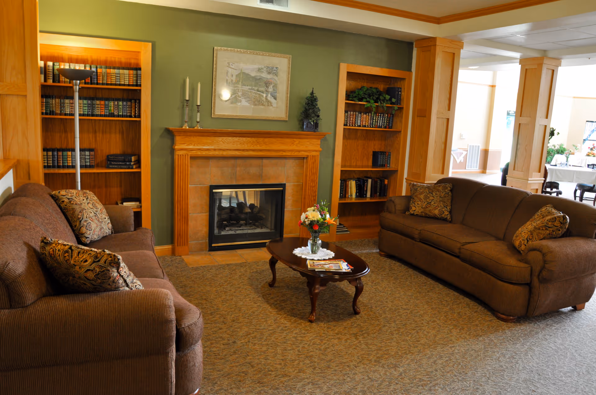 A cozy living room area in a senior living facility featuring two brown upholstered sofas with patterned cushions facing each other across a wooden coffee table with a vase of flowers and magazines. The room has a fireplace with a wooden mantle, flanked by built-in wooden bookshelves filled with books and decorative items. The walls are painted green with a framed picture above the fireplace, and the space is warmly lit with natural light coming from an adjacent room.