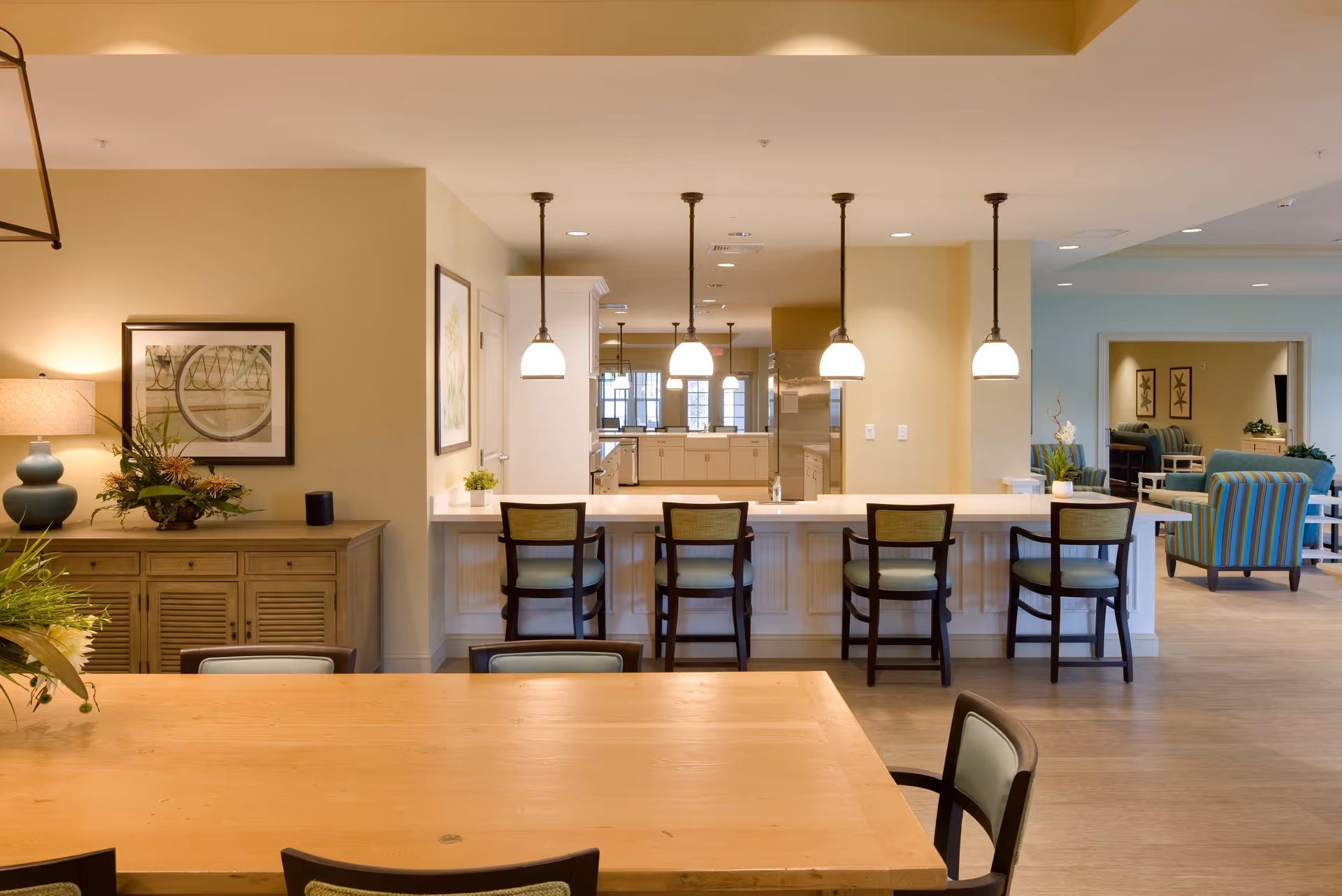 Open communal dining and kitchen area with a long wooden table in the foreground, bar seating with pendant lights, and lounge seating in the background.