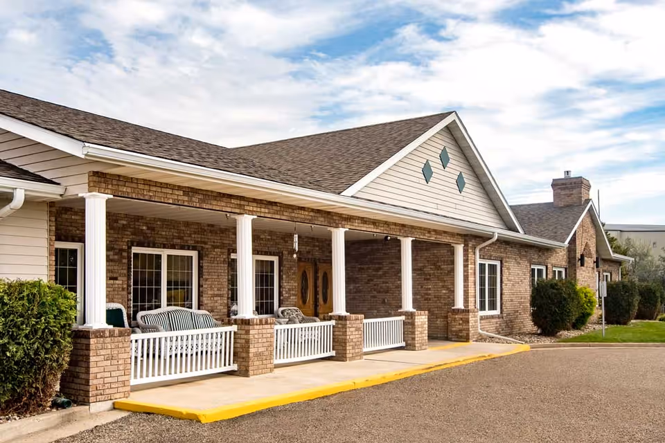 Front exterior of a brick senior living building with a covered porch, white columns, and outdoor seating.