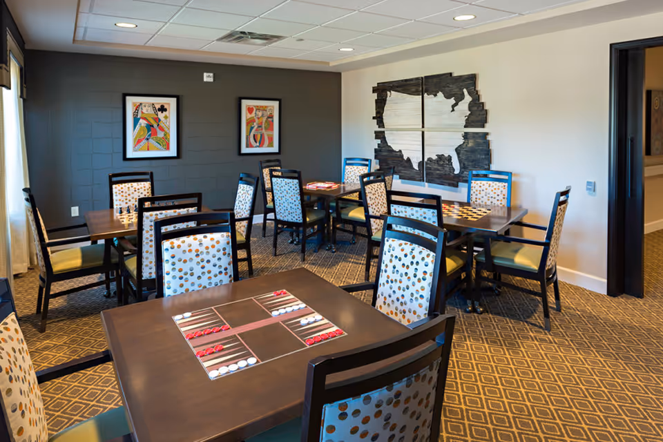 A well-lit room with several tables and chairs arranged for board games. The tables have built-in game boards with red and white game pieces. The chairs have patterned upholstery with circular designs. The walls are decorated with framed artwork, including two playing card images and a large wooden map. The carpet has a diamond pattern, and the ceiling has recessed lighting.