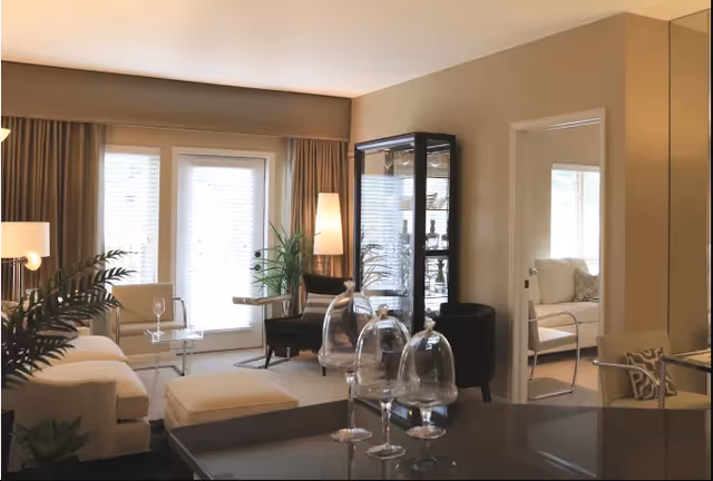 A cozy living room with beige walls and large windows covered by curtains. The room features a white sofa, a matching ottoman, a glass coffee table, and two black armchairs. There is a tall floor lamp and a glass display cabinet with decorative items. In the foreground, there are several empty wine glasses on a dark countertop. A doorway leads to another room with a white chair and cushions.