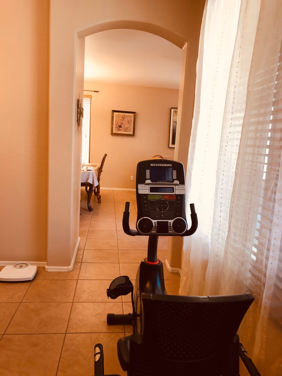 An exercise bike sits on a tiled hallway near a window with sheer curtains, with a dining room visible through an arched doorway.