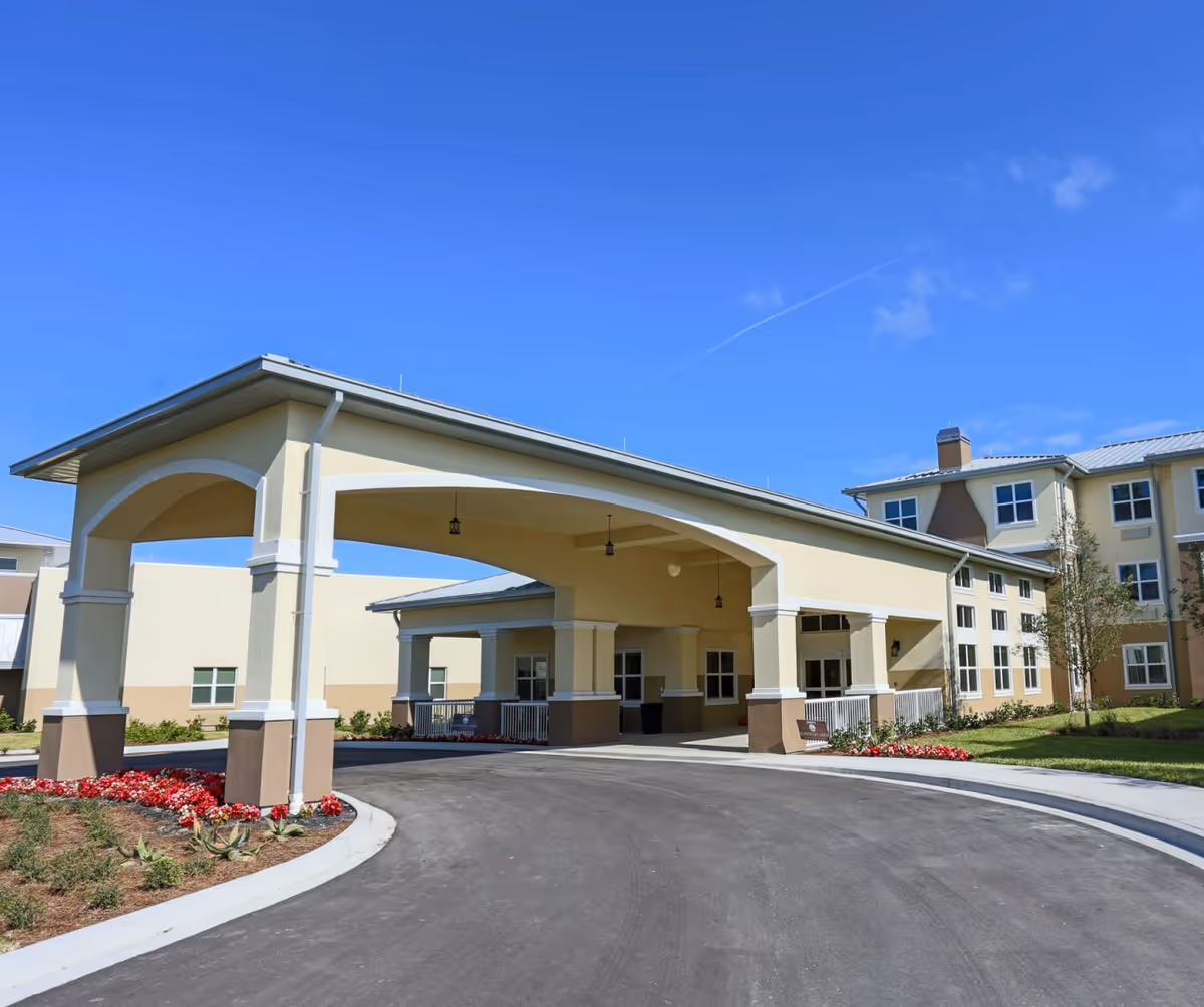 Covered porte-cochère entrance of a multi-story beige assisted living building with landscaping under a clear blue sky.