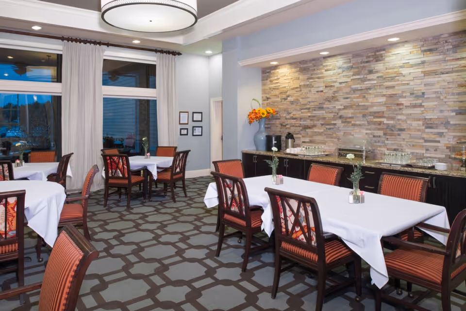 Dining room with tables covered in white tablecloths, red-upholstered chairs, patterned carpet, and a stone accent wall with a buffet.
