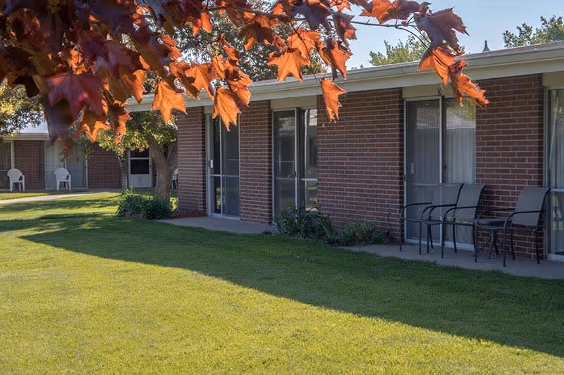 Exterior view of a single-story brick building with sliding glass doors leading to small patios furnished with chairs and tables. The foreground features a well-maintained green lawn and a tree with orange autumn leaves partially framing the top of the image.