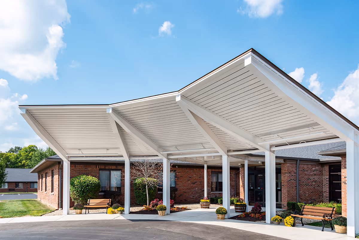 Covered entrance canopy with benches and potted plants at the front of a brick senior living building under a blue sky.