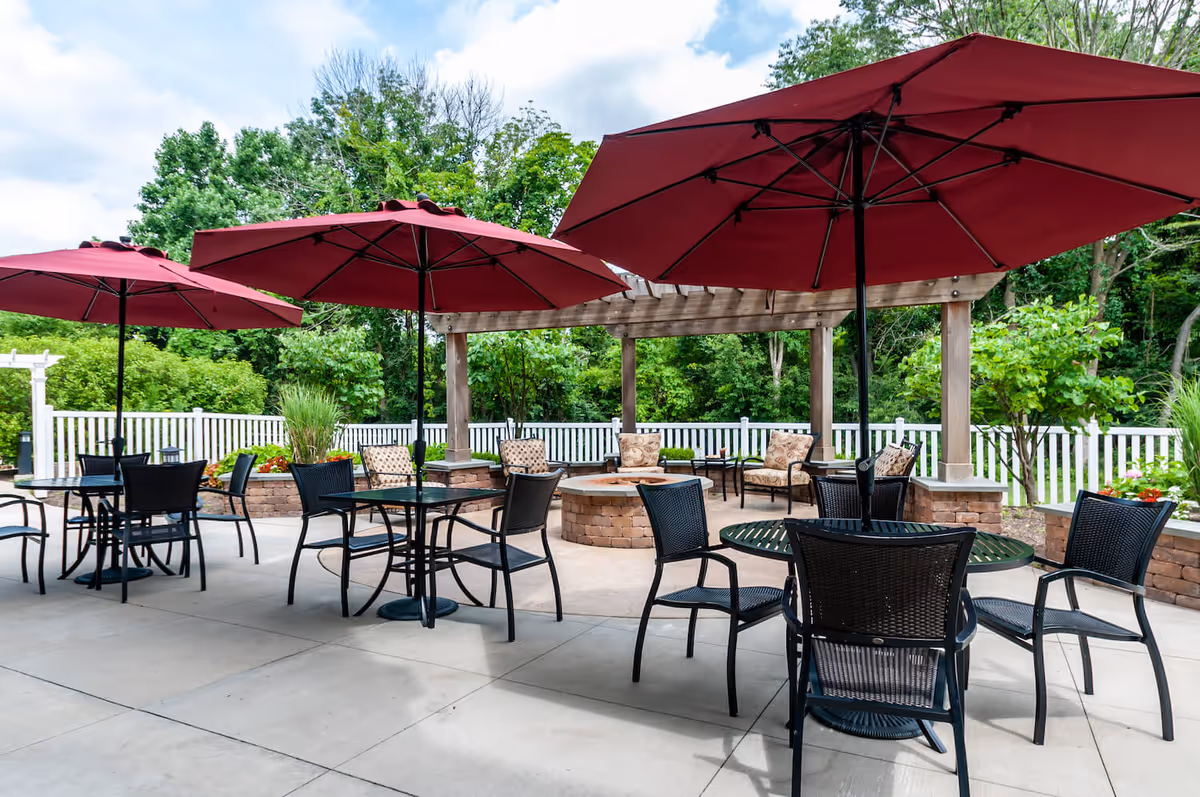 Outdoor patio with tables, chairs, red umbrellas, a pergola and seating area surrounded by greenery.