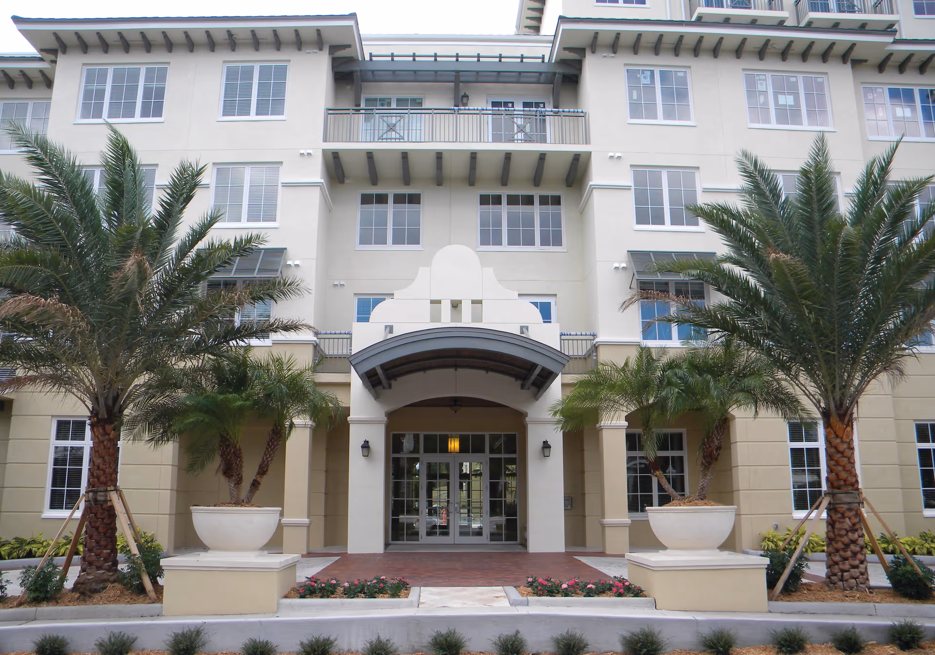 Front entrance of a multi-story senior living building with palm trees and large planters flanking a covered entryway.