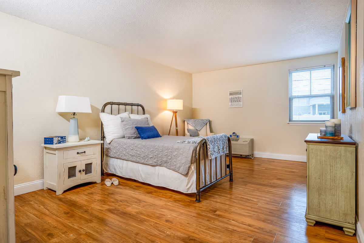 A cozy bedroom with a metal frame bed dressed in gray and white bedding with blue accent pillows. There is a wooden nightstand with a lamp and a box of tissues on the left side of the bed. A pair of slippers is placed on the wooden floor near the nightstand. The room has light-colored walls, a window with blinds, a small dresser with candles on the right, and a framed picture on the wall.