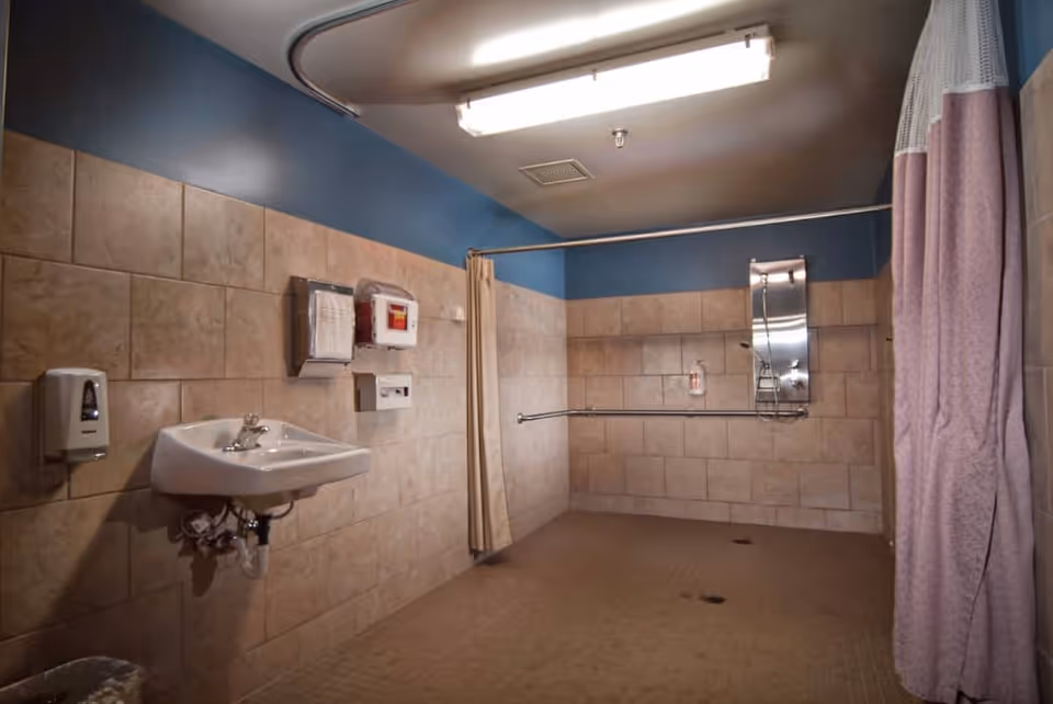 A spacious tiled bathroom with beige tiles on the walls and floor, a white wall-mounted sink with a soap dispenser, a paper towel dispenser, a red emergency box, a mirror, grab bars, and a shower area with a curtain.