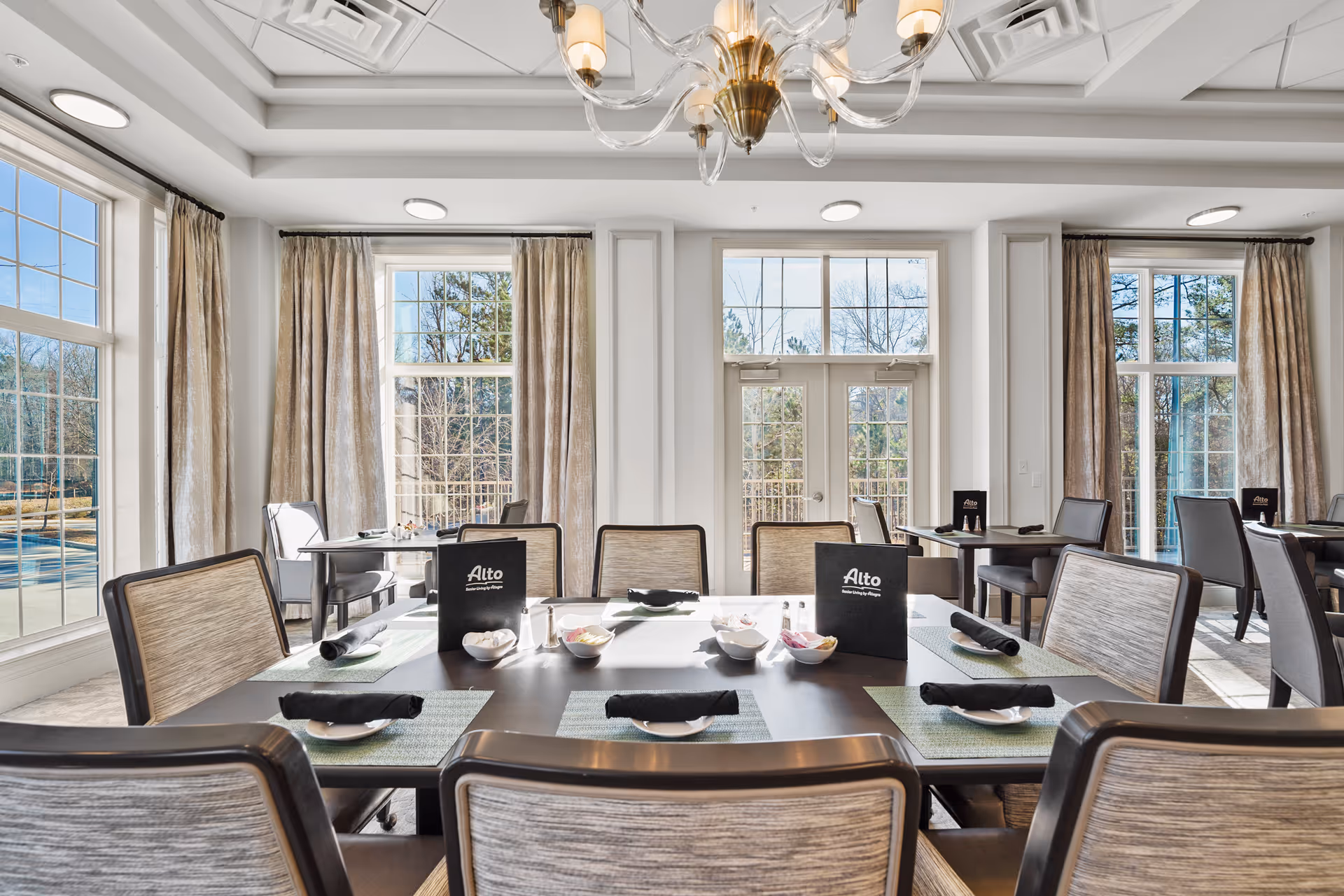 Bright dining room with large windows and beige curtains letting in natural light. Several tables are set with black napkins, small bowls, and Alto Johns Creek menus. The room features a chandelier and light-colored walls with decorative molding.
