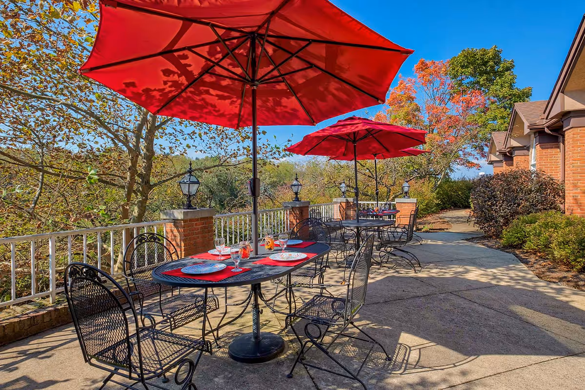 Outdoor patio with wrought-iron tables and chairs under red umbrellas, place settings, and a brick building beside a tree-lined view.