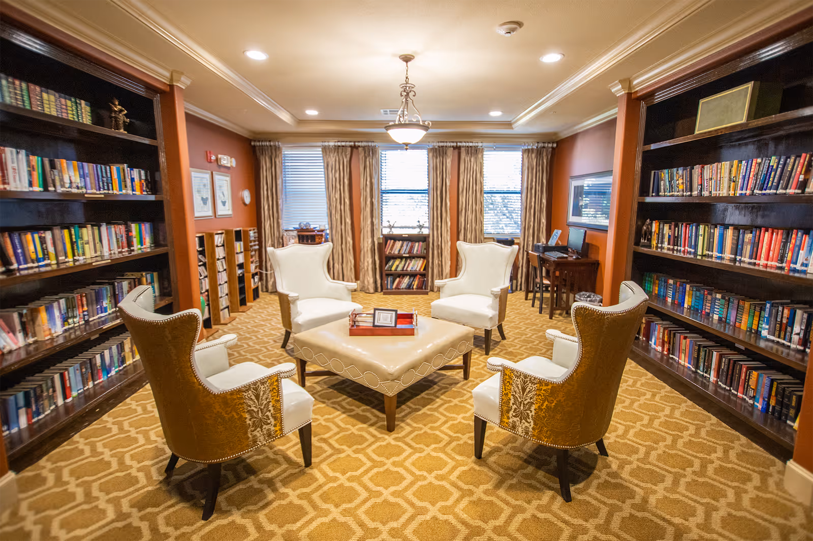 A cozy library room with four upholstered armchairs arranged around a square ottoman in the center. The room has built-in dark wood bookshelves filled with books on both sides, a patterned carpet, and large windows with curtains letting in natural light. There is a small desk with a computer in the back corner.