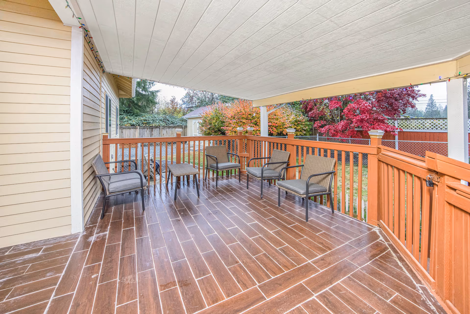 Covered wood‑look deck with several chairs and a small table overlooking a fenced backyard with autumn trees.