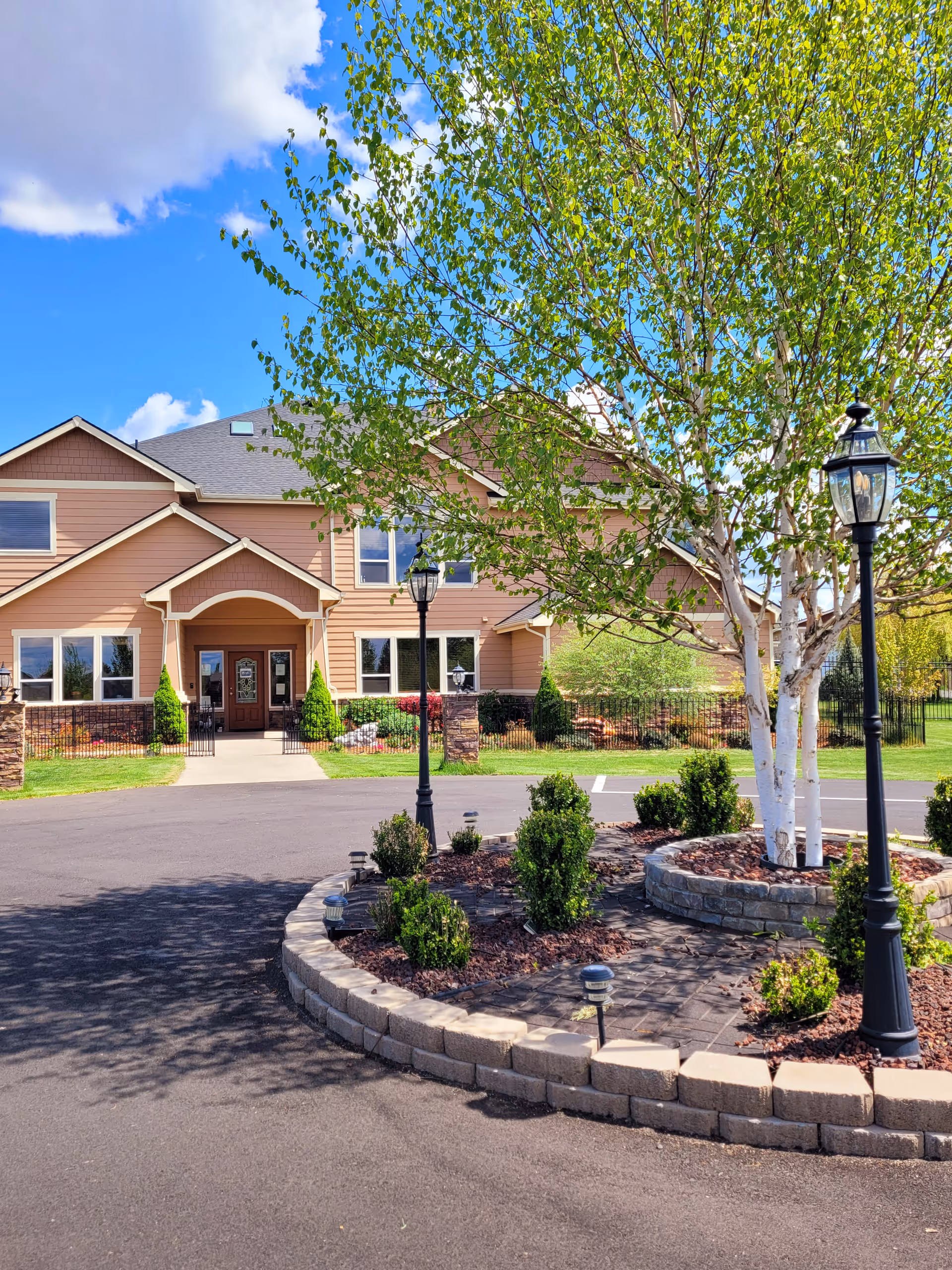 Front exterior of a two-story senior living facility with a landscaped circular planter, birch trees, and lampposts in the driveway.