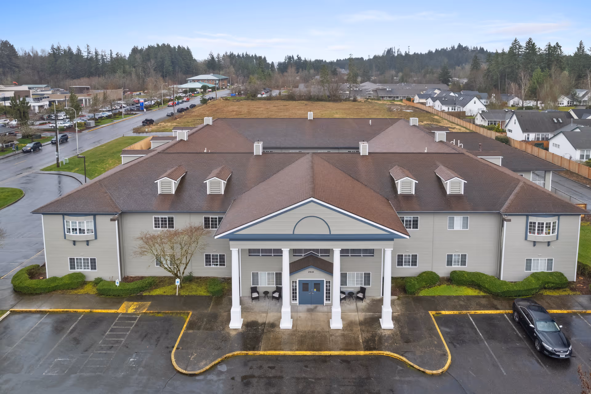Aerial front view of a two-story assisted living building with a columned main entrance, parking lot, and surrounding neighborhood.